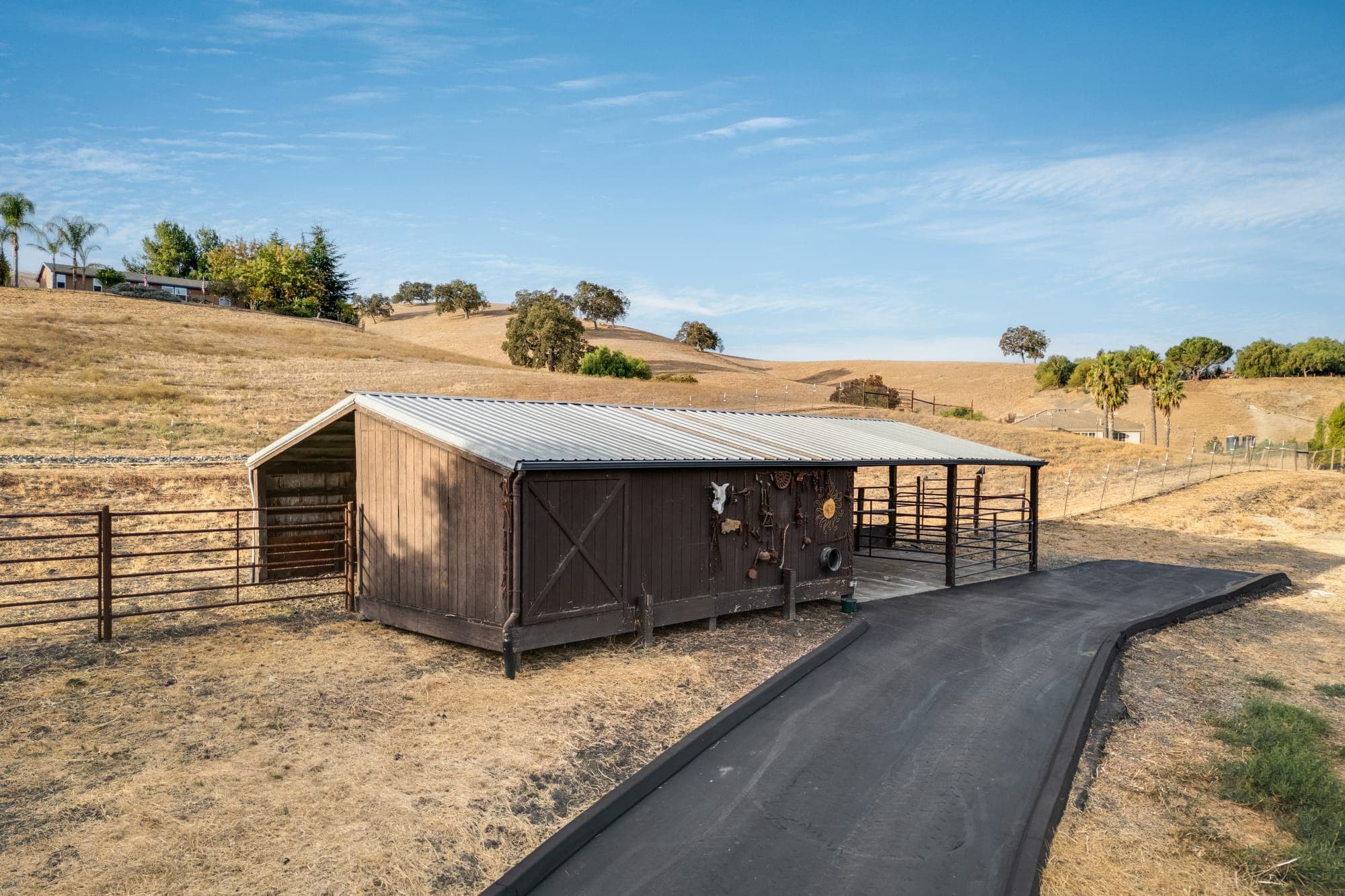 The covered one-stall barn with dark wood siding, a metal roof, and a covered run, beside a paved path.