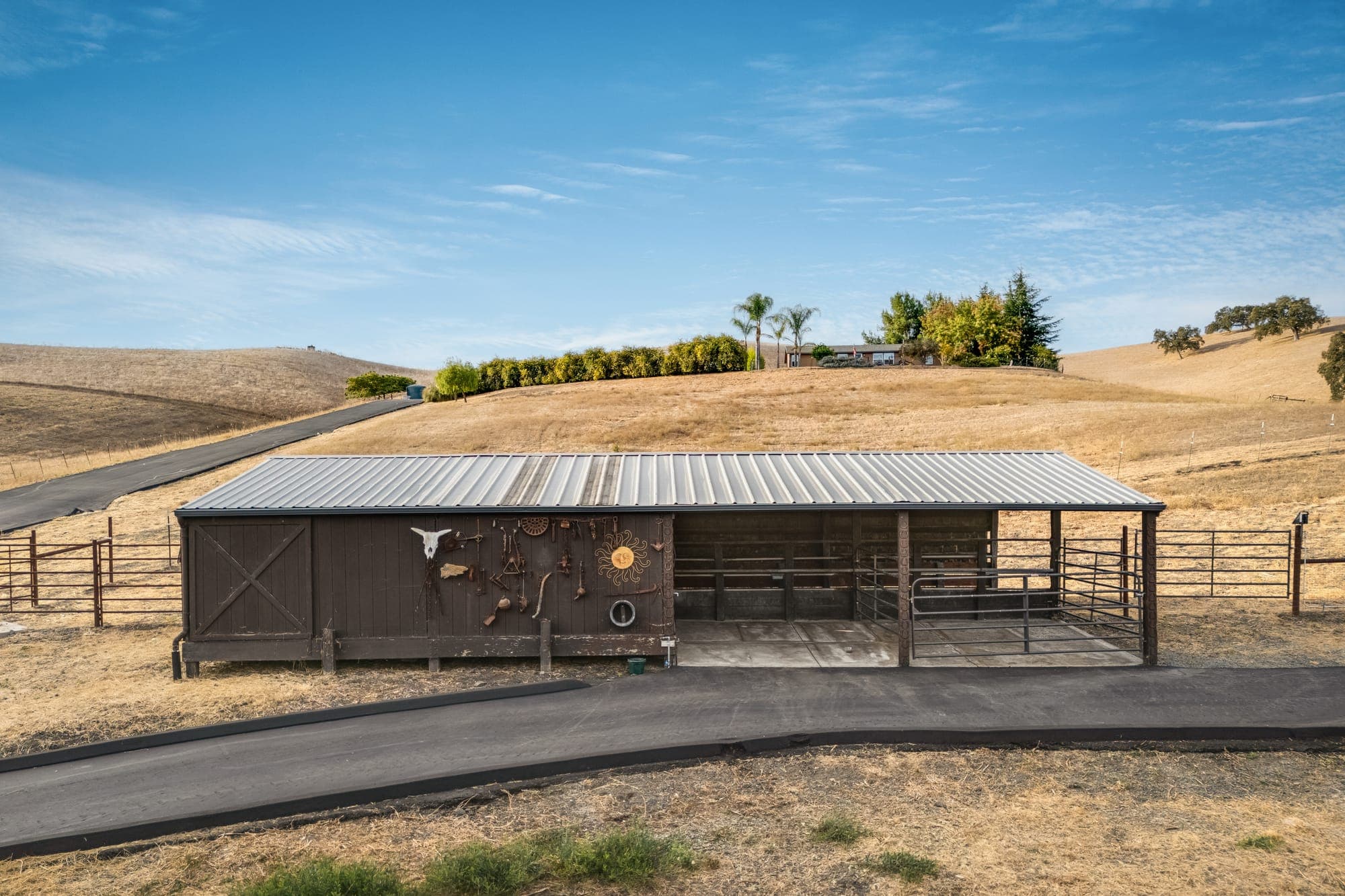 The barn from the front, with wood siding, a covered run, and pipe-panel fencing against the hills.
