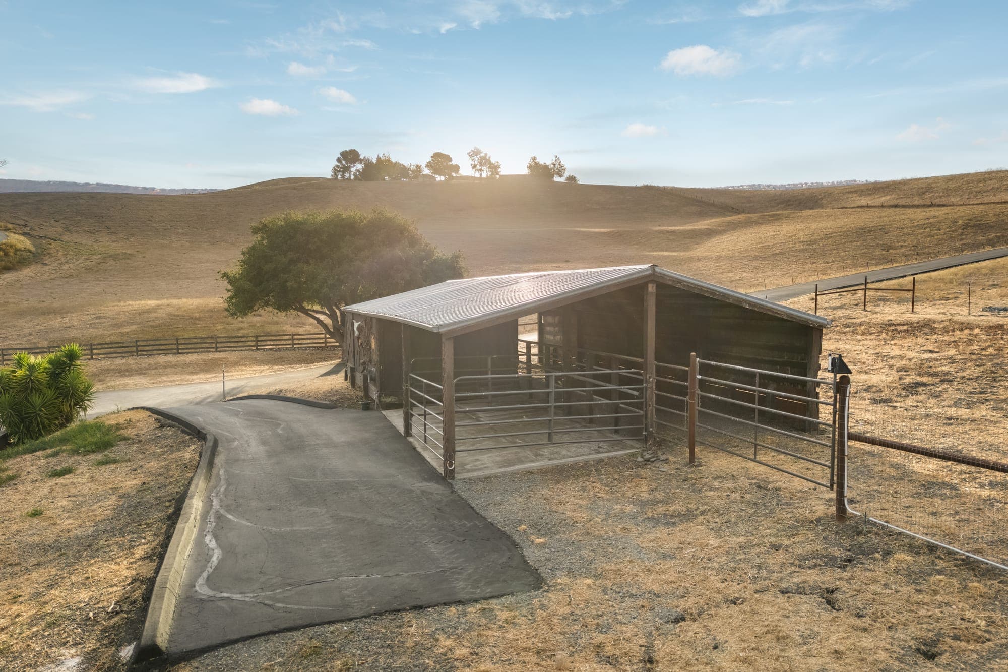 A covered shelter with a pipe-panel pen at the end of a paved path, oaks on the hill behind.