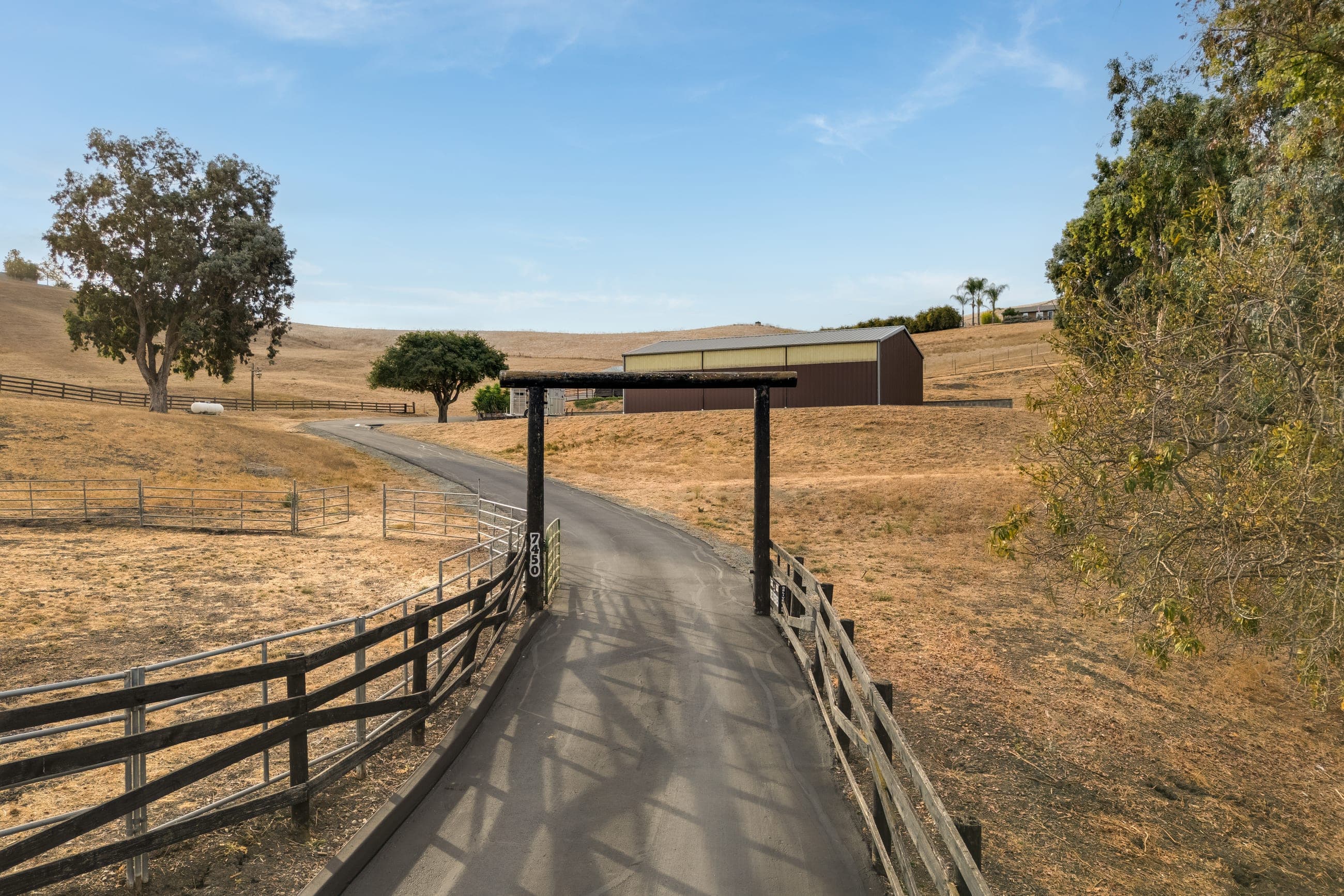 The timber entry gate and paved drive at 7450 Johnston Road, between wood rail fences, with golden hills and the shop beyond.