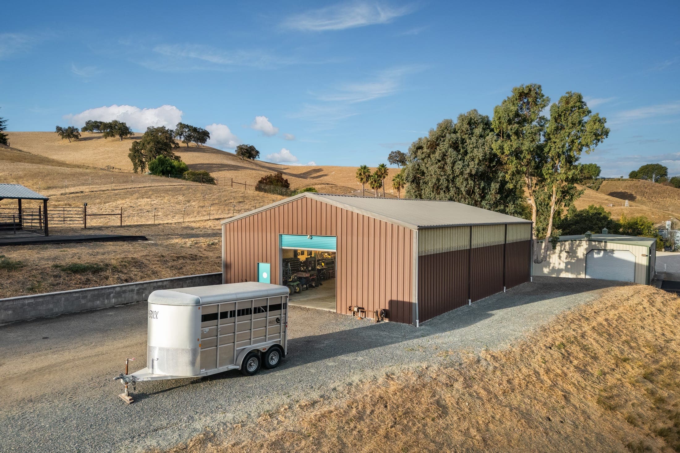 The 2,400 sq ft shop with its roll-up doors, a horse trailer on the apron, and hills beyond.